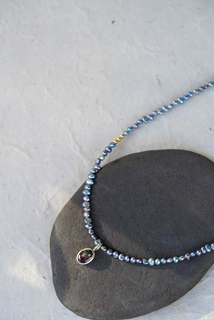 A delicate beaded necklace with small iridescent blue-gray pearls, gold spacer accents, and a tourmaline at the center, laid on a light textured surface.
