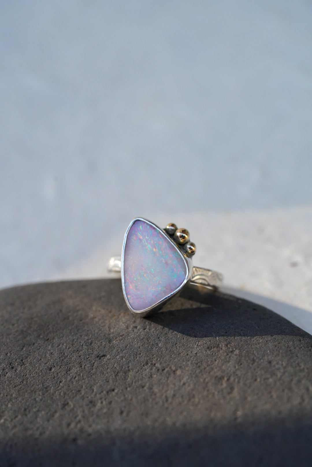 A triangular iridescent opal ring with a silver stamped band with gold accents on the stone, resting on a dark textured stone against a light gray background.