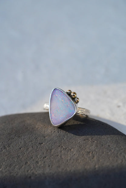 A triangular iridescent opal ring with a silver stamped band with gold accents on the stone, resting on a dark textured stone against a light gray background.