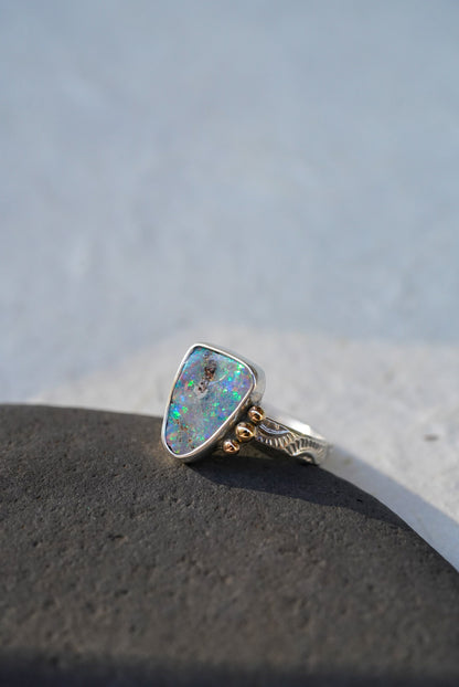 A silver ring with a triangular iridescent opal gemstone and small gold decorative accents on the band, resting on a dark textured surface against a light gray background.