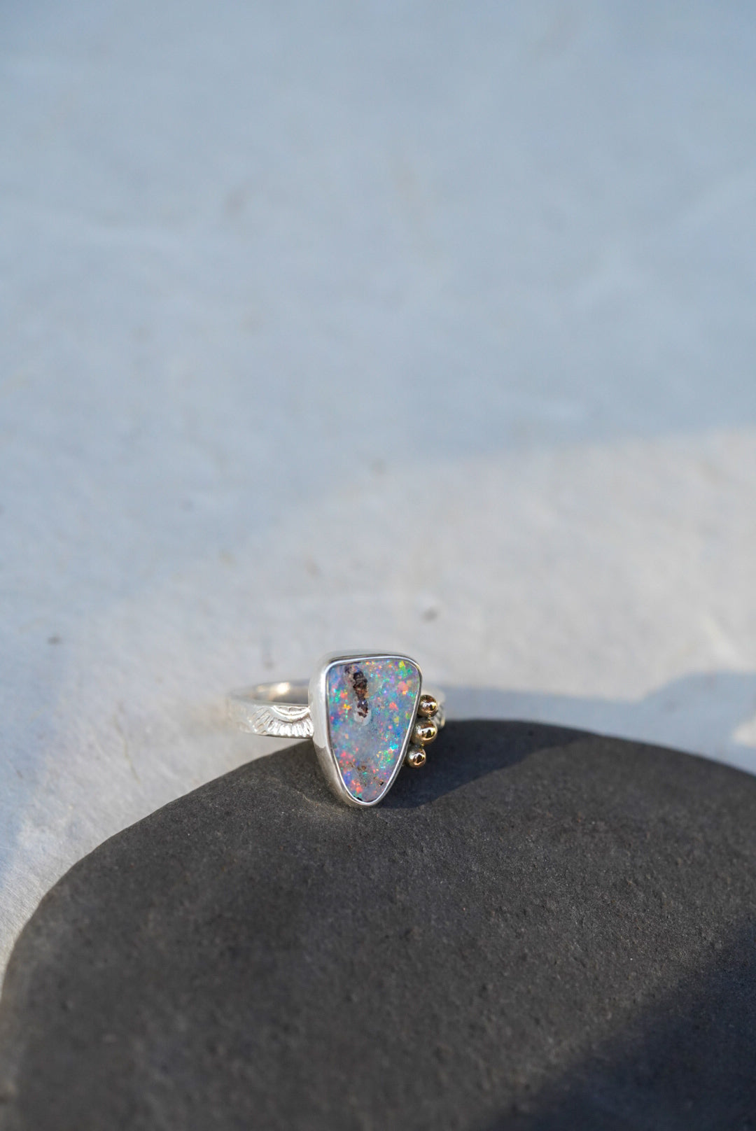 A silver ring with a triangular iridescent opal gemstone and small gold decorative accents on the band, resting on a dark textured surface against a light gray background.