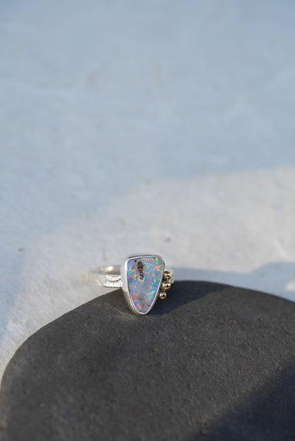A silver ring with a triangular iridescent opal gemstone and small gold decorative accents on the band, resting on a dark textured surface against a light gray background.