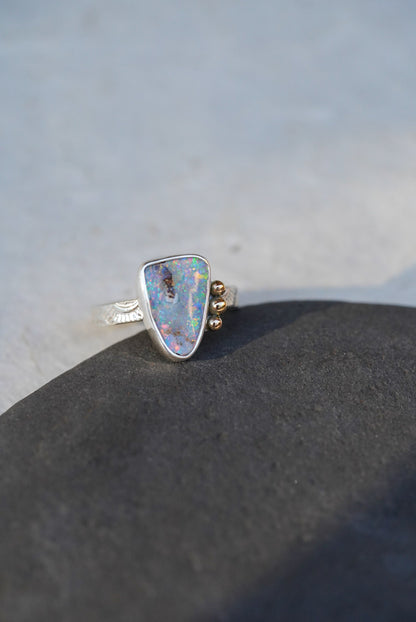 A silver ring with a triangular iridescent opal gemstone and small gold decorative accents on the band, resting on a dark textured surface against a light gray background.