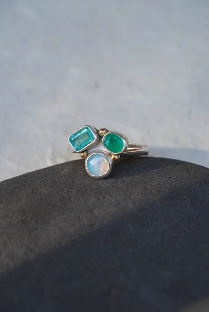A silver ring with three gemstones—a rectangular emerald-cut emerald, a round emerald stone, and a round white opal—resting on a dark textured surface against a light gray background.