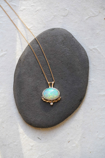 A gold necklace with an oval iridescent opal pendant with a twisted gold accent and small diamond below, resting on a dark gray stone against a textured off-white background.