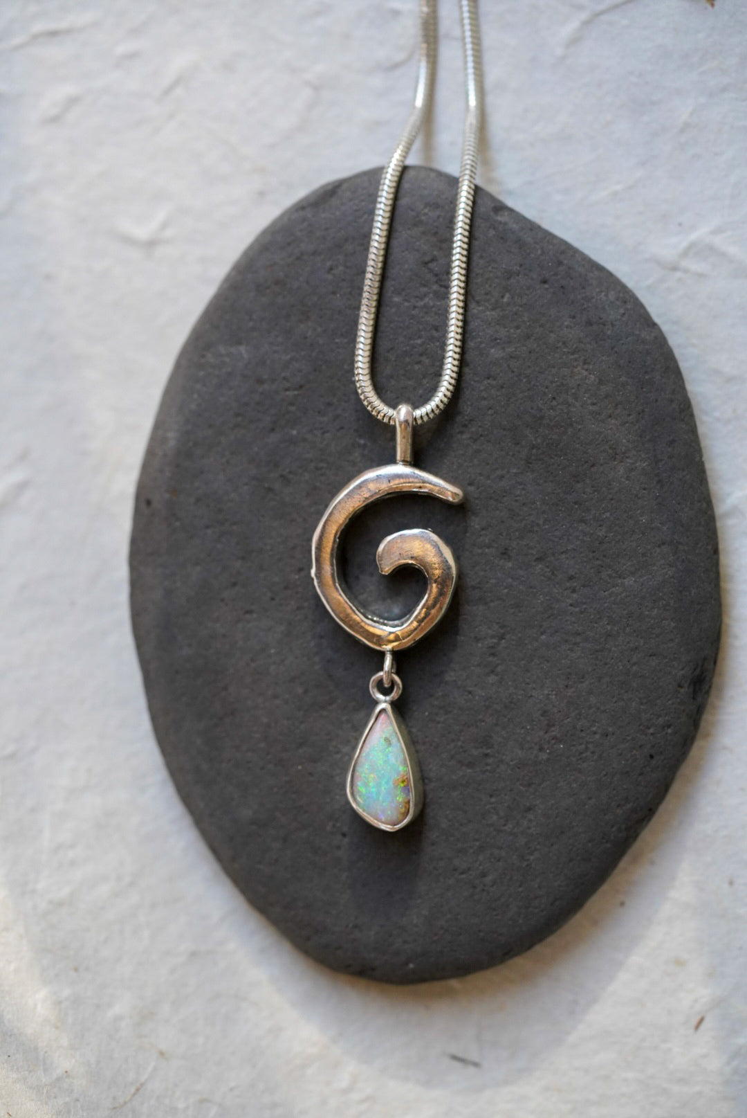 A silver necklace with a textured spiral pendant and a teardrop-shaped opal stone dangle, displayed on a dark gray stone against a light textured background.