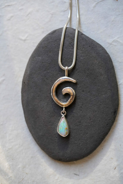 A silver necklace with a textured spiral pendant and a teardrop-shaped opal stone dangle, displayed on a dark gray stone against a light textured background.