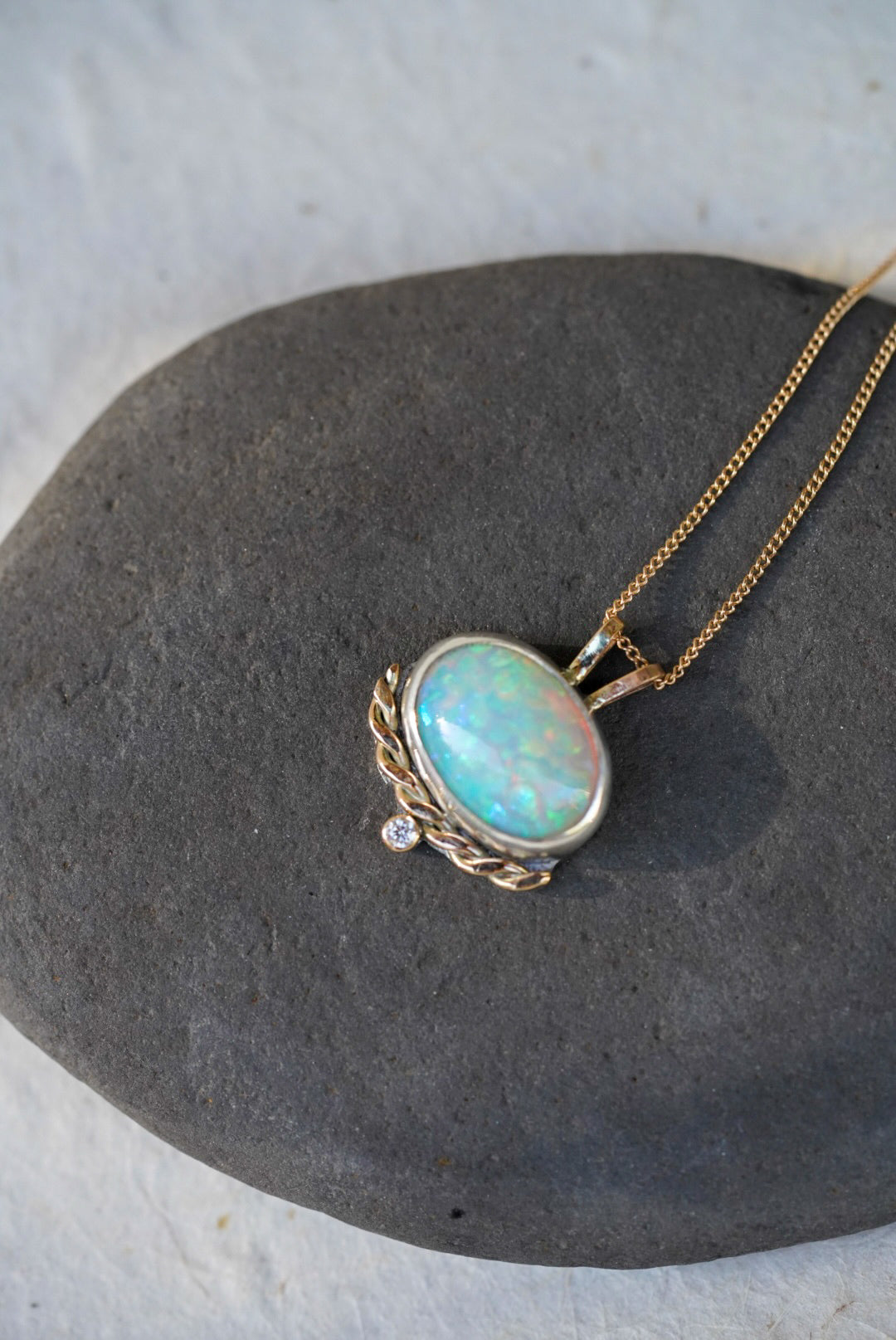 A gold necklace with an oval iridescent opal pendant with a twisted gold accent and small diamond below, resting on a dark gray stone against a textured off-white background.