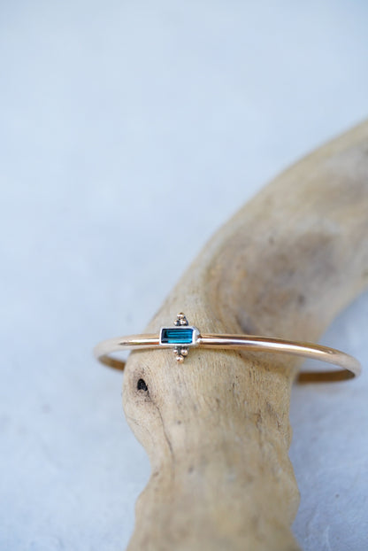 A delicate gold cuff featuring a small rectangular blue-green gemstone, displayed on a curved piece of driftwood against a light textured background.