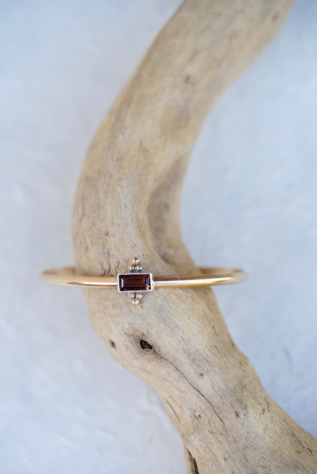 A delicate gold cuff featuring a small rectangular brown/purple gemstone, displayed on a curved piece of driftwood against a light textured background.
