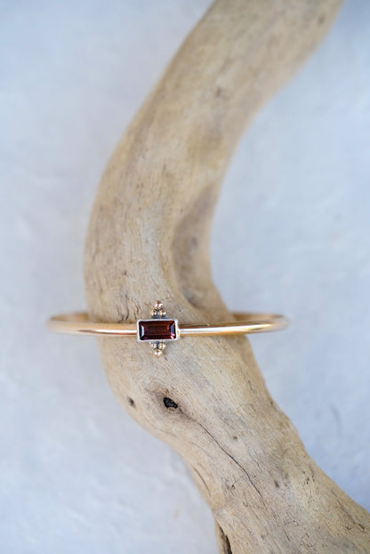A delicate gold cuff featuring a small rectangular brown/purple gemstone, displayed on a curved piece of driftwood against a light textured background.