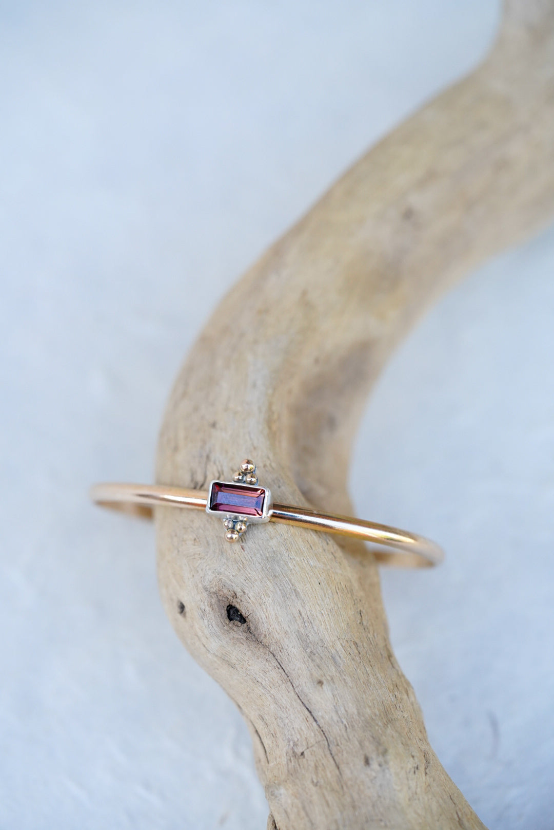 A delicate gold cuff featuring a small rectangular brown/purple gemstone, displayed on a curved piece of driftwood against a light textured background.