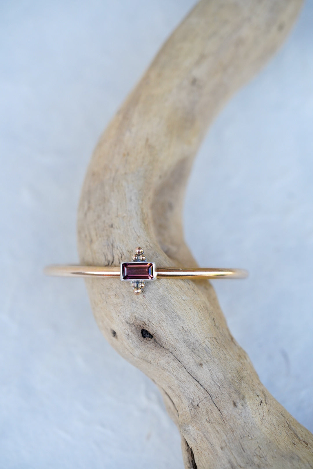 A delicate gold cuff featuring a small rectangular brown/purple gemstone, displayed on a curved piece of driftwood against a light textured background.