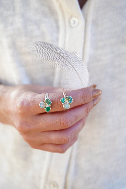 A close-up of a person's hand holding a delicate white feather, wearing two rings set with green and blue stones, against a soft cream-colored linen shirt.