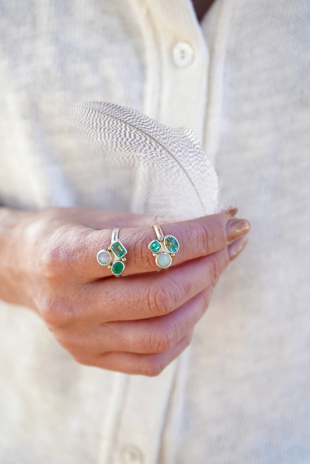 A close-up of a person's hand holding a delicate white feather, wearing two rings with green and pale blue stones, against a soft cream-colored linen shirt.