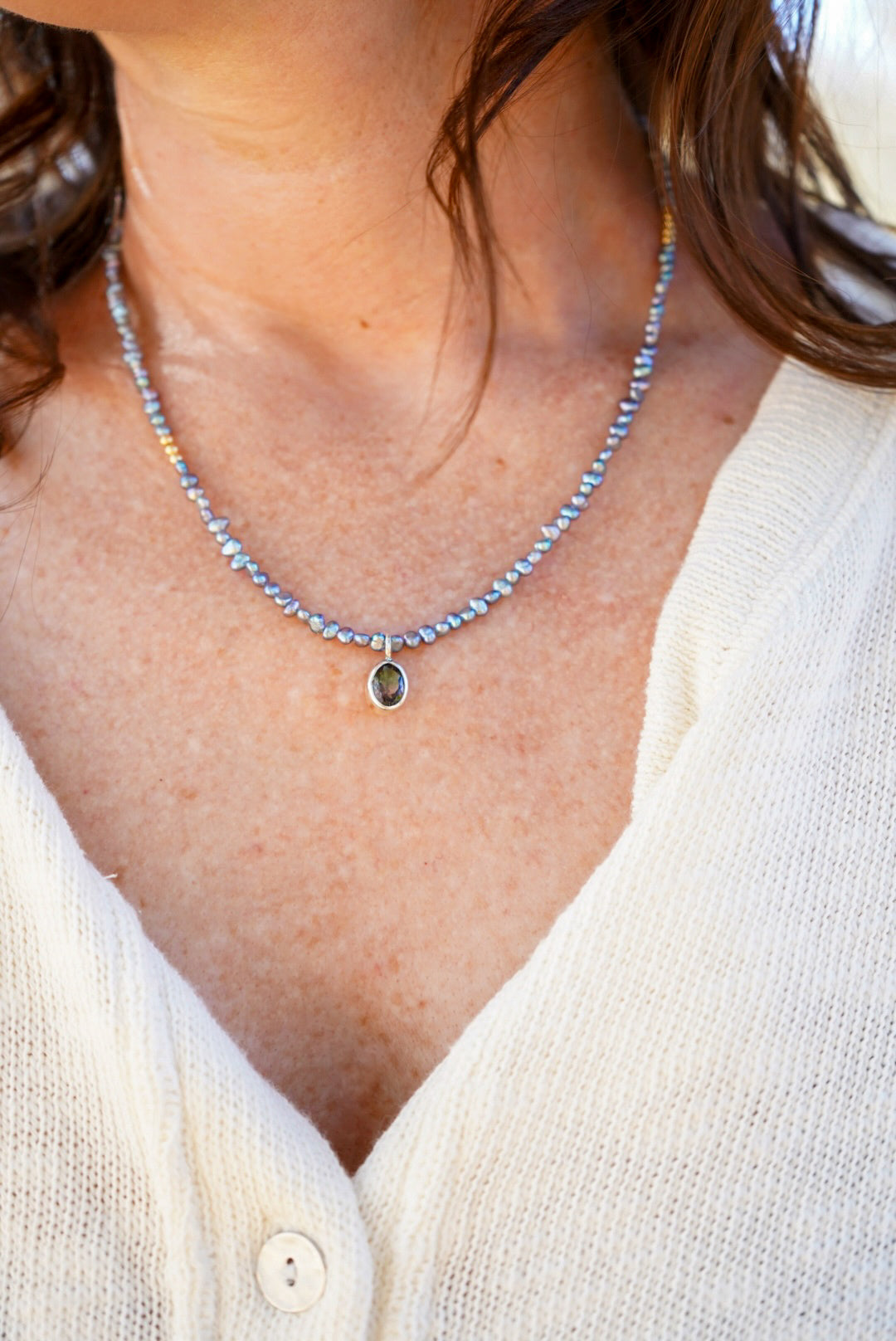 A close-up of a woman wearing a delicate pearl beaded necklace with a small oval tourmaline, paired with a cream-colored knit top.