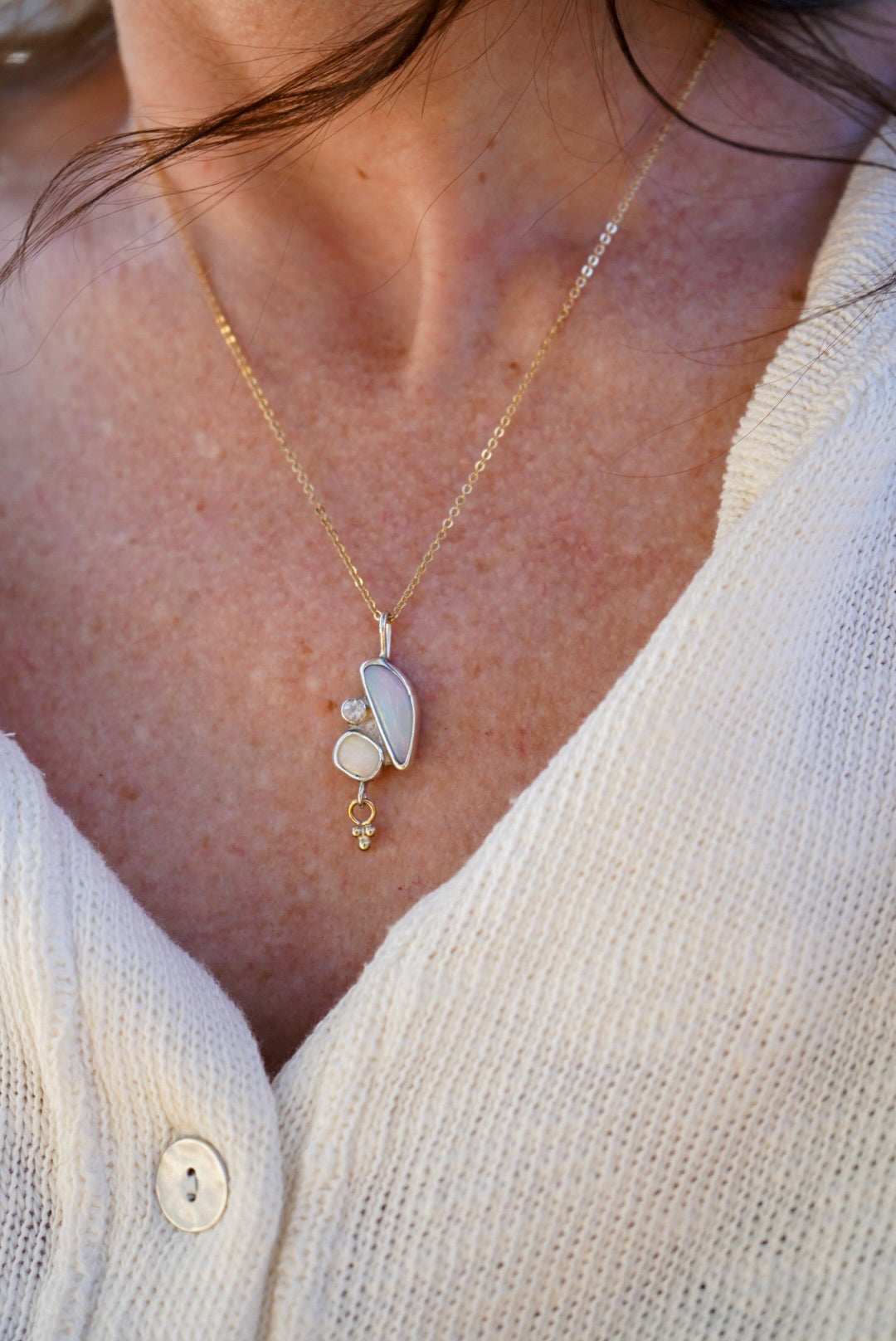 A close-up of a person wearing a delicate gold necklace with a pendant made of opals and a small moonstone with a gold charm hanging below, paired with a white knit cardigan.