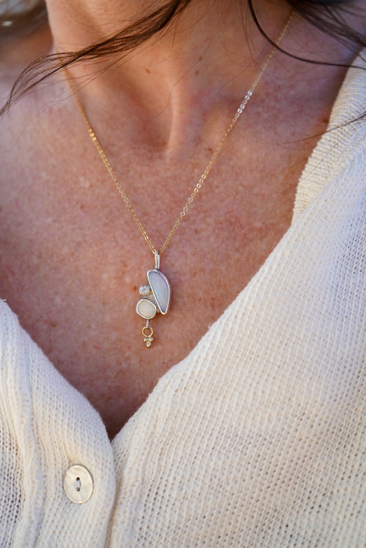 A close-up of a person wearing a delicate gold necklace with a pendant made of opals and a small moonstone with a gold charm hanging below, paired with a white knit cardigan.