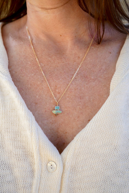 A close-up of a person wearing a delicate gold chain necklace with a small pendant featuring a light blue-green tourmaline stone and opal with a tiny gold accent, styled with a cream-colored knit cardigan.