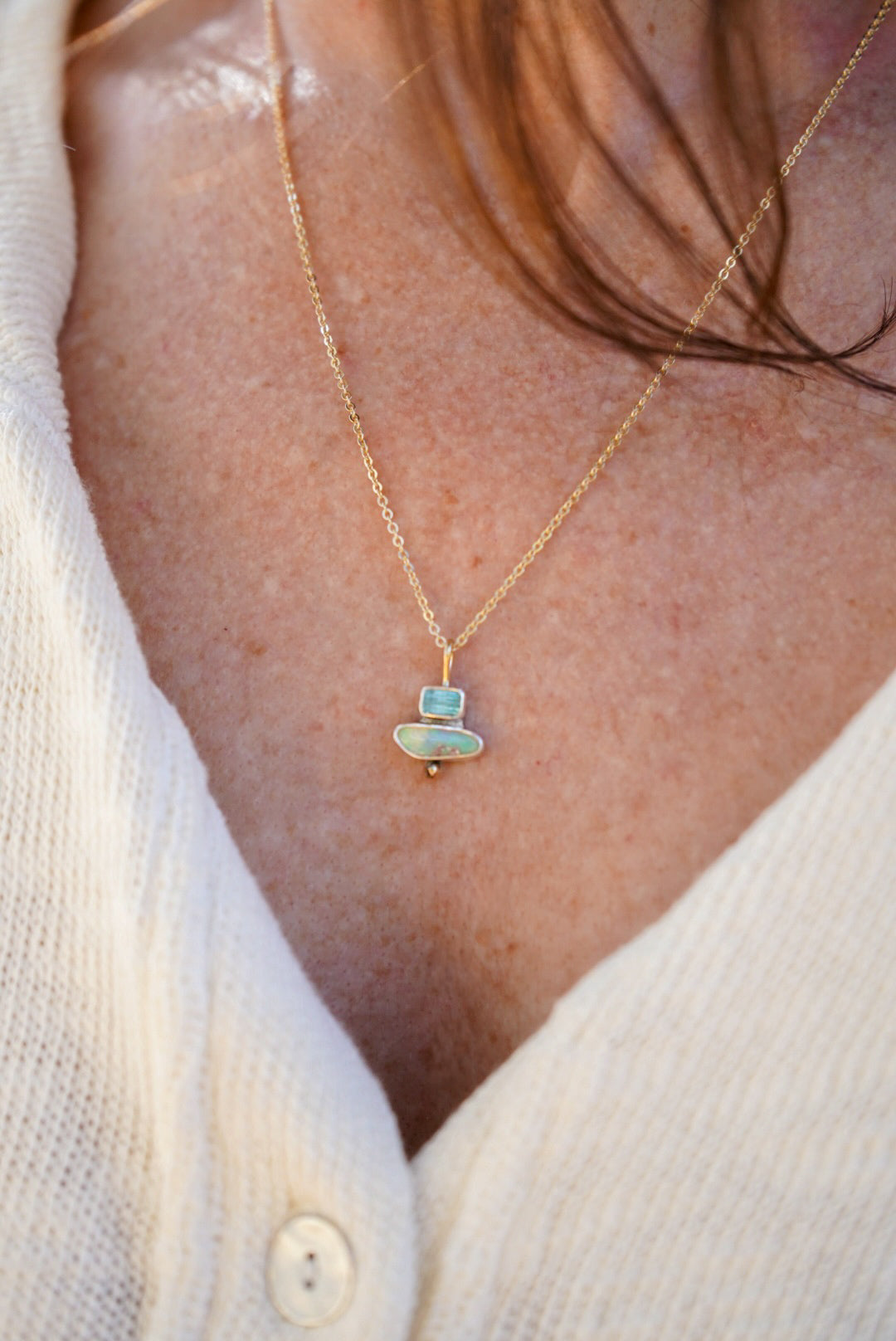 A close-up of a person wearing a delicate gold chain necklace with a small pendant featuring a light blue-green tourmaline stone and opal with a tiny gold accent, styled with a cream-colored knit cardigan.