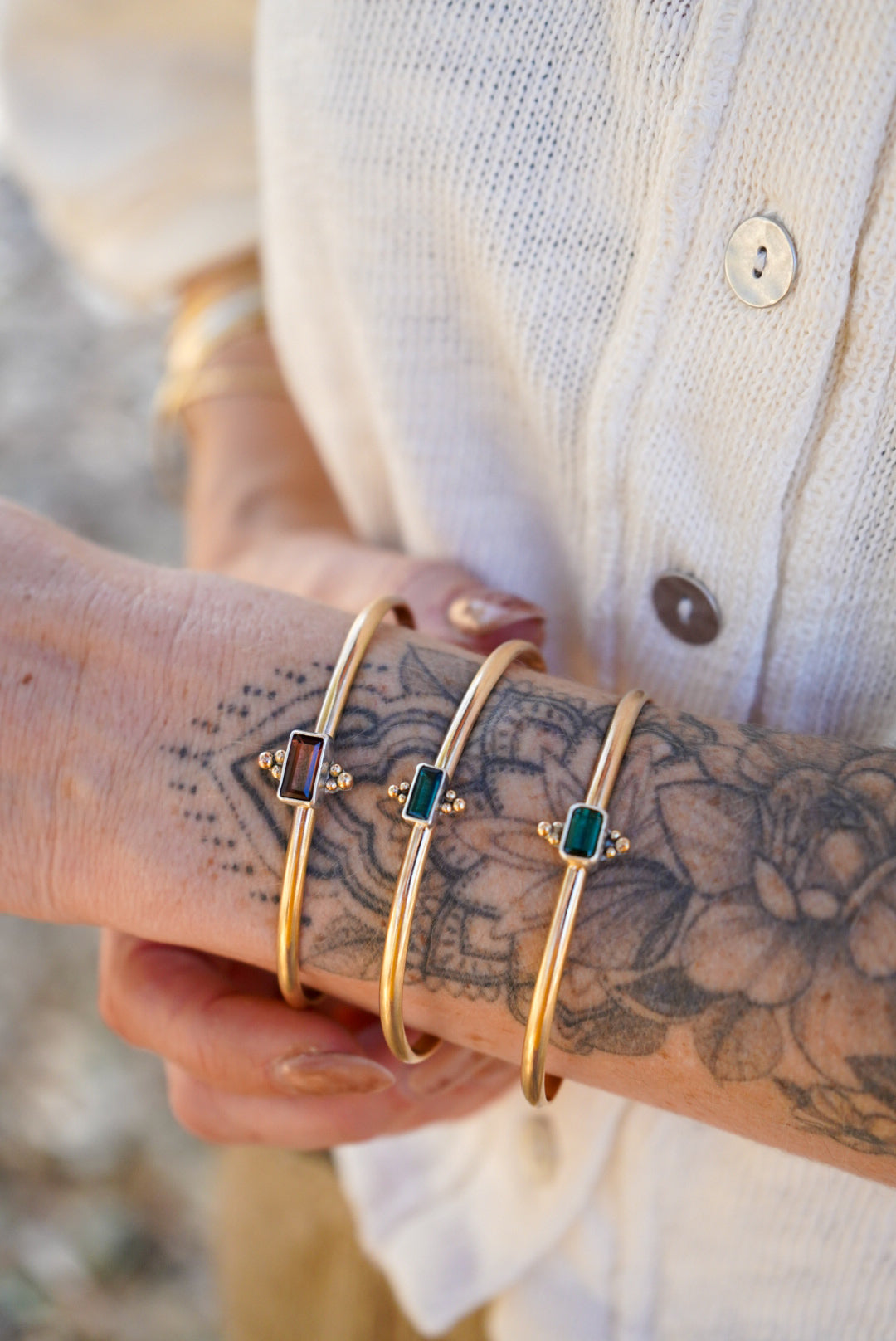 Three gold bangle bracelets with rectangular colored tourmaline stones are shown worn on a person's wrist, with a white button-up shirt visible in the background.