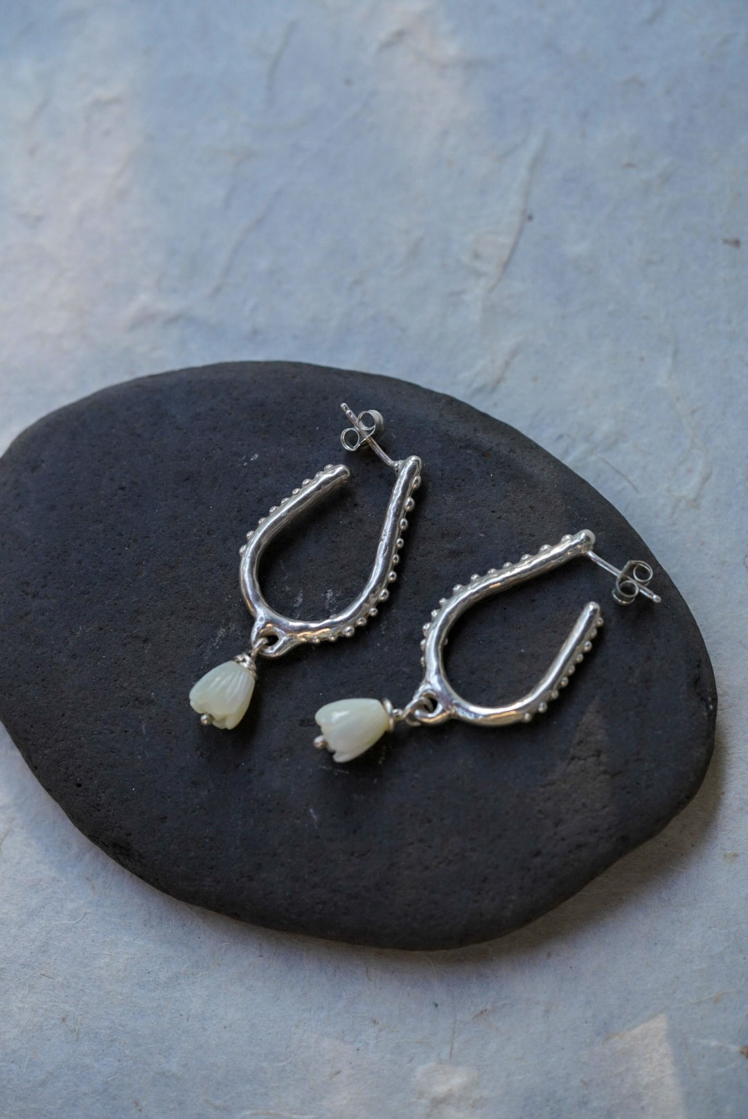 A pair of silver hoop earrings with hand carved flower beads, displayed on a dark stone against a light gray textured background.
