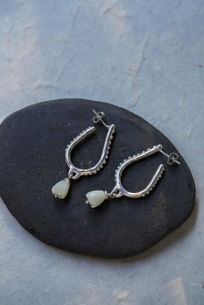 A pair of silver hoop earrings with hand carved flower beads, displayed on a dark stone against a light gray textured background.