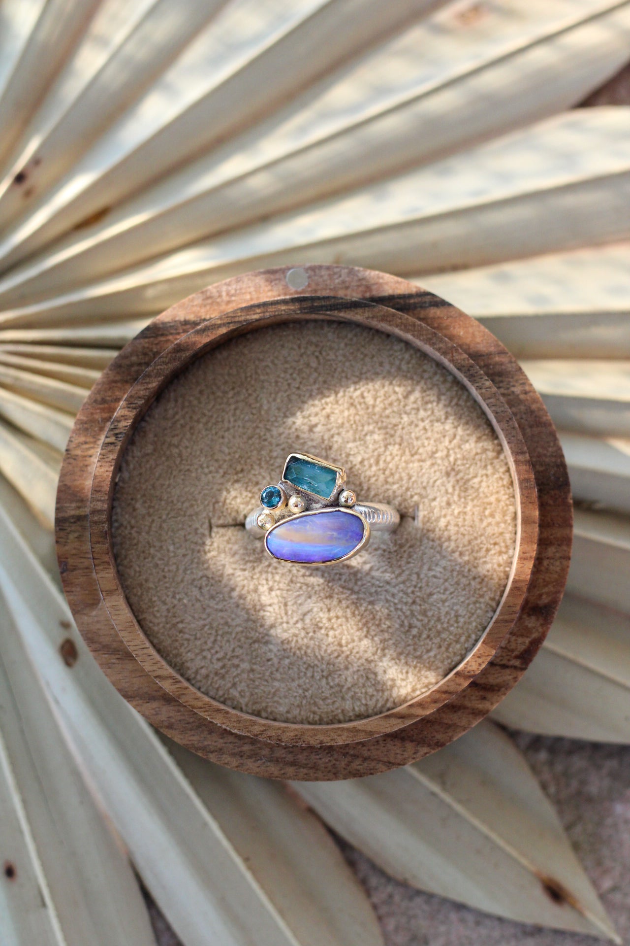 Ring with an opal, tourmaline and blue topaz on a wooden display box with sand, surrounded by dried palm leaves.