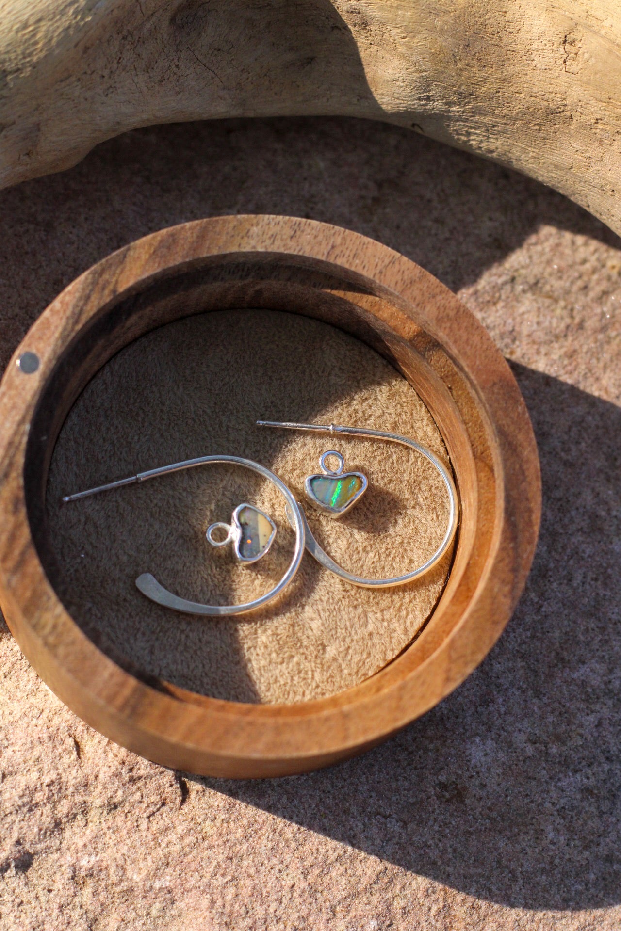 Silver hoop earrings with heart shaped opal charms in a wooden box on a stone surface