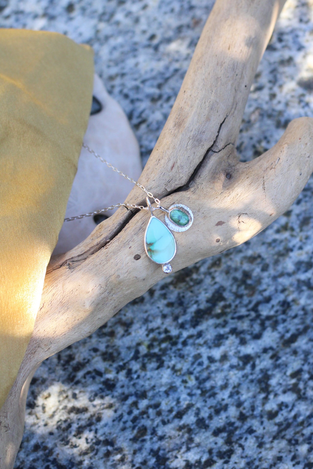 Turquoise teardrop pendant on a chain draped over a piece of driftwood on a beach.