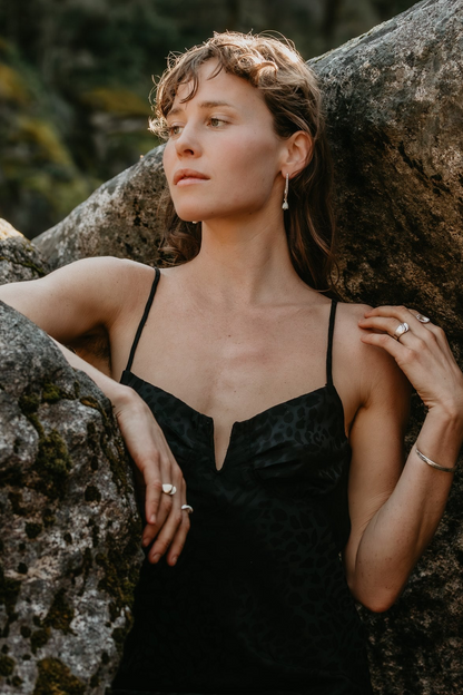 A woman with curly light brown hair leans against a large mossy rock, wearing a black leopard-print spaghetti-strap camisole top and silver jewelry.