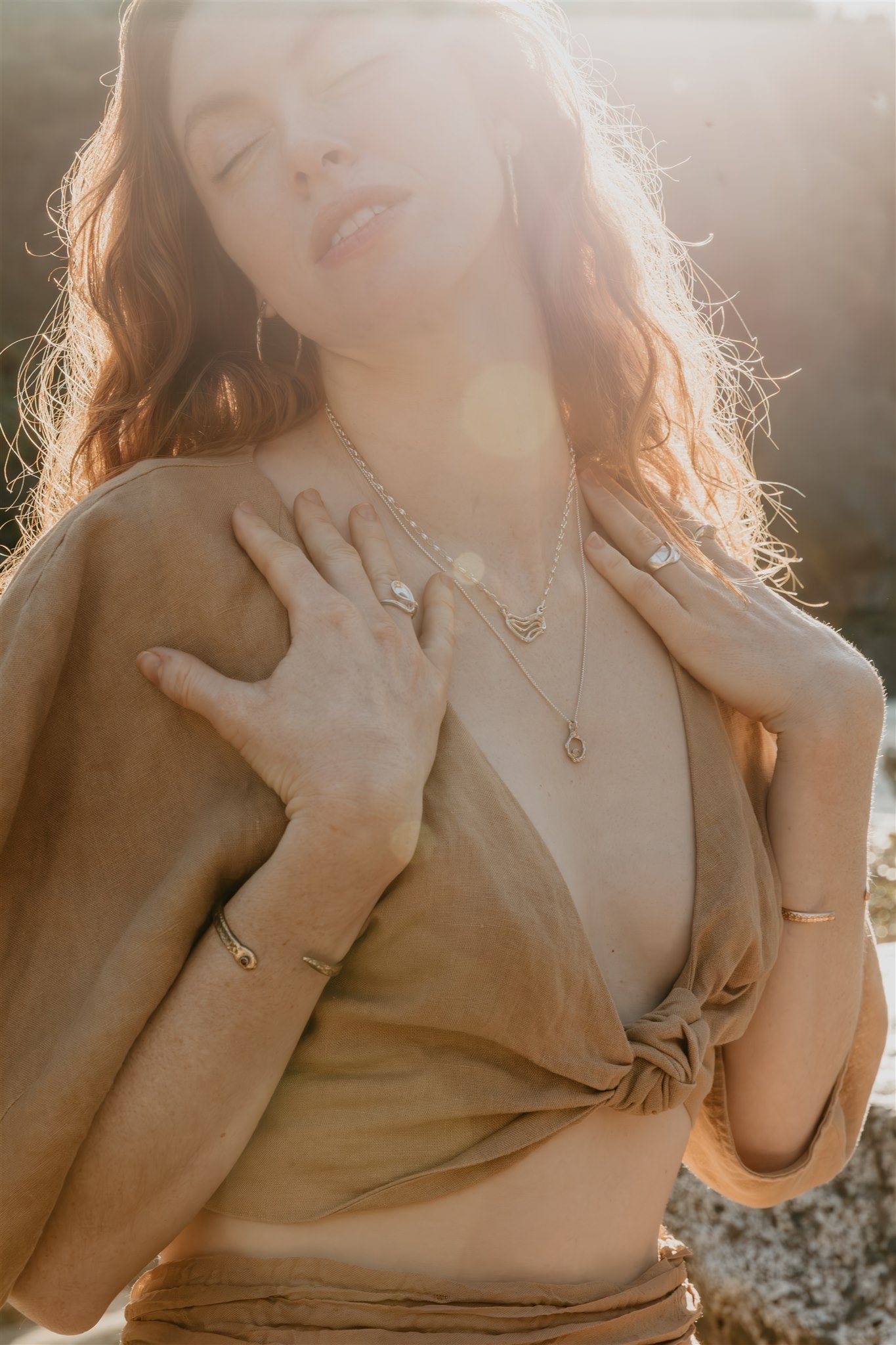 Woman with closed eyes wearing a beige top and gold jewelry against a blurred natural background