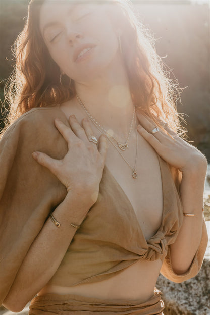 Woman with closed eyes wearing a beige top and gold jewelry against a blurred natural background