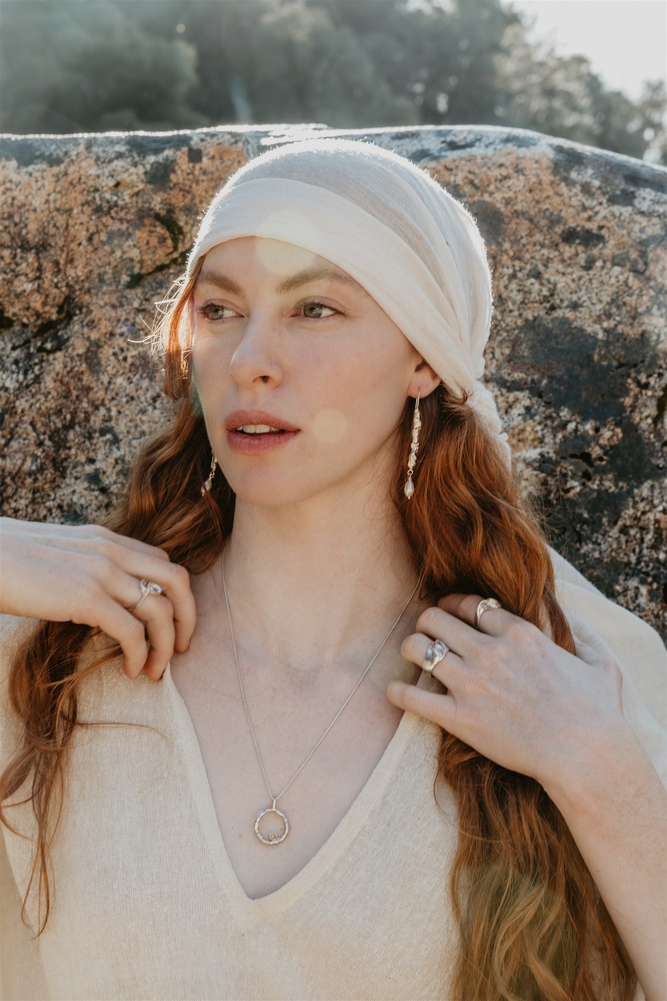 Woman with long hair wearing a white headscarf and beige sweater against a stone wall, wearing jewelry