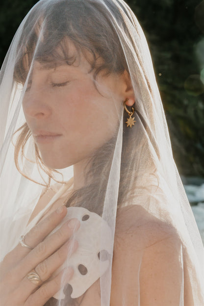 Woman wearing a veil with a blurred natural background, wearing rings and a star earring.