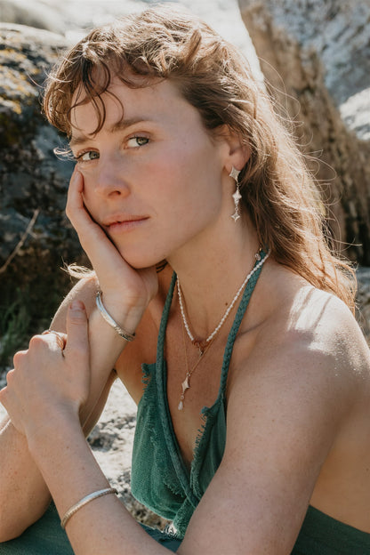 Woman with long hair and green top sitting outdoors near rocks wearing jewelry
