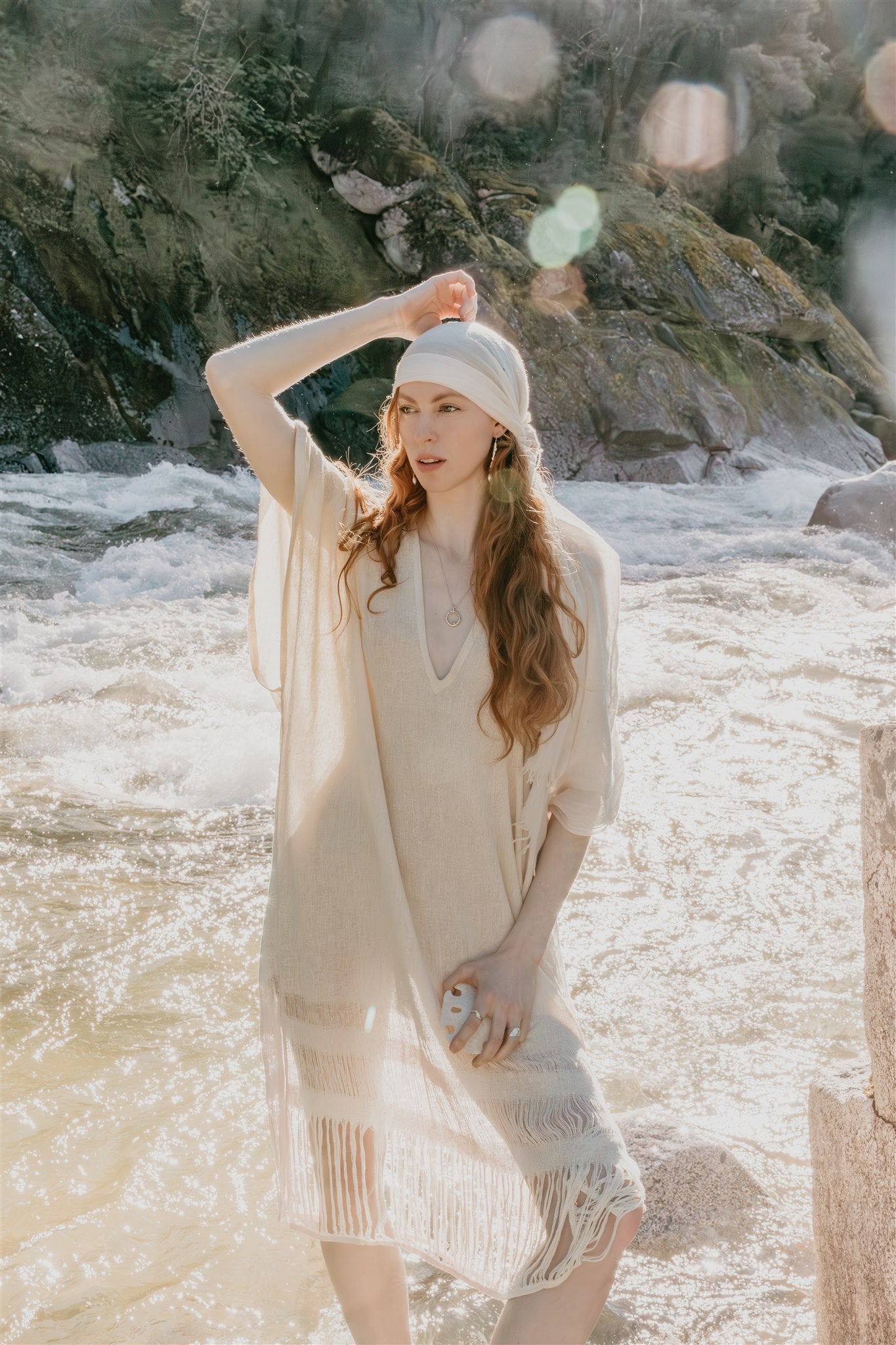 Woman in a beige dress standing by a flowing river with rocks and greenery.