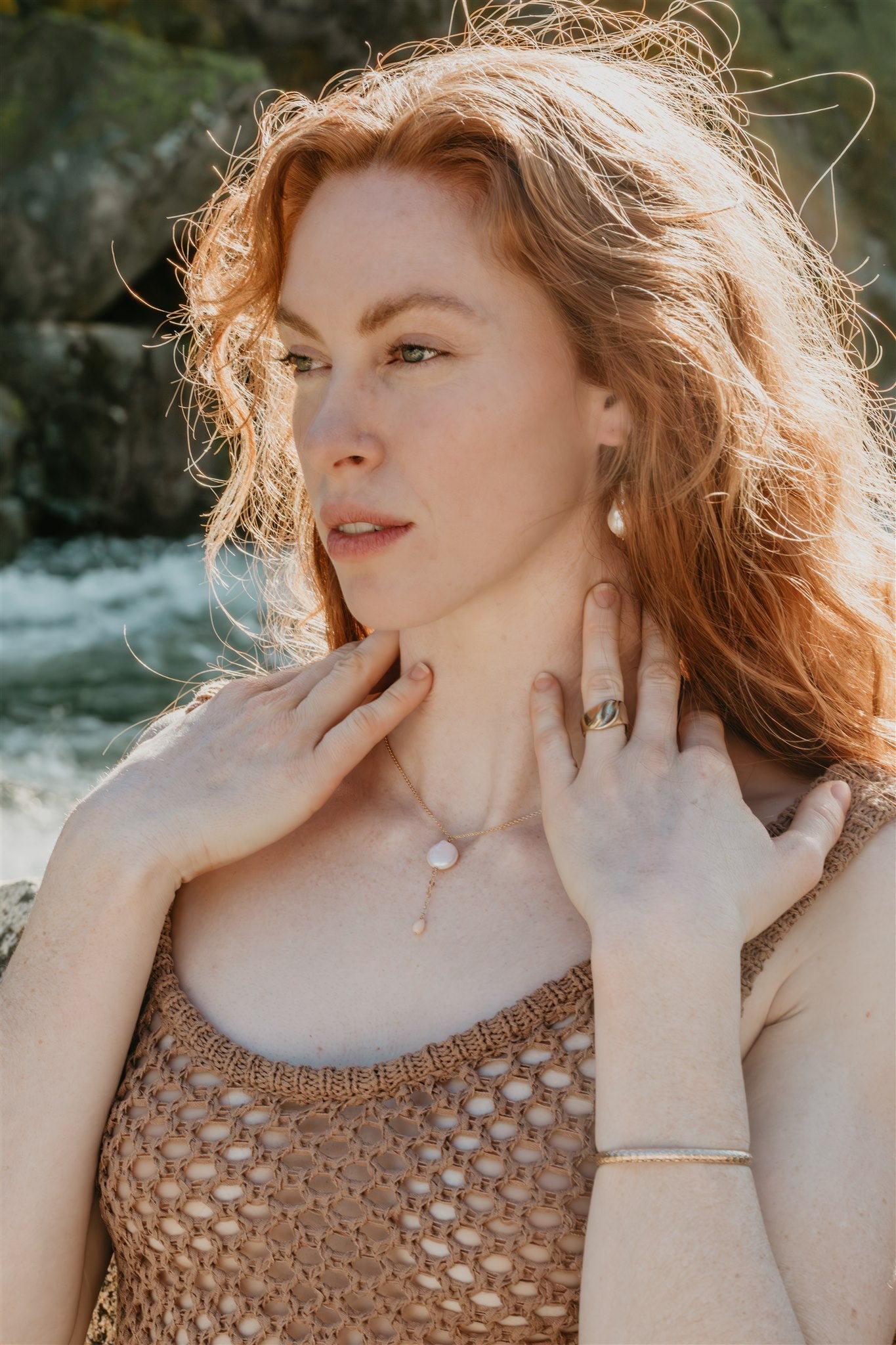 Woman touching her neck, wearing a necklace and ring, with a natural background.