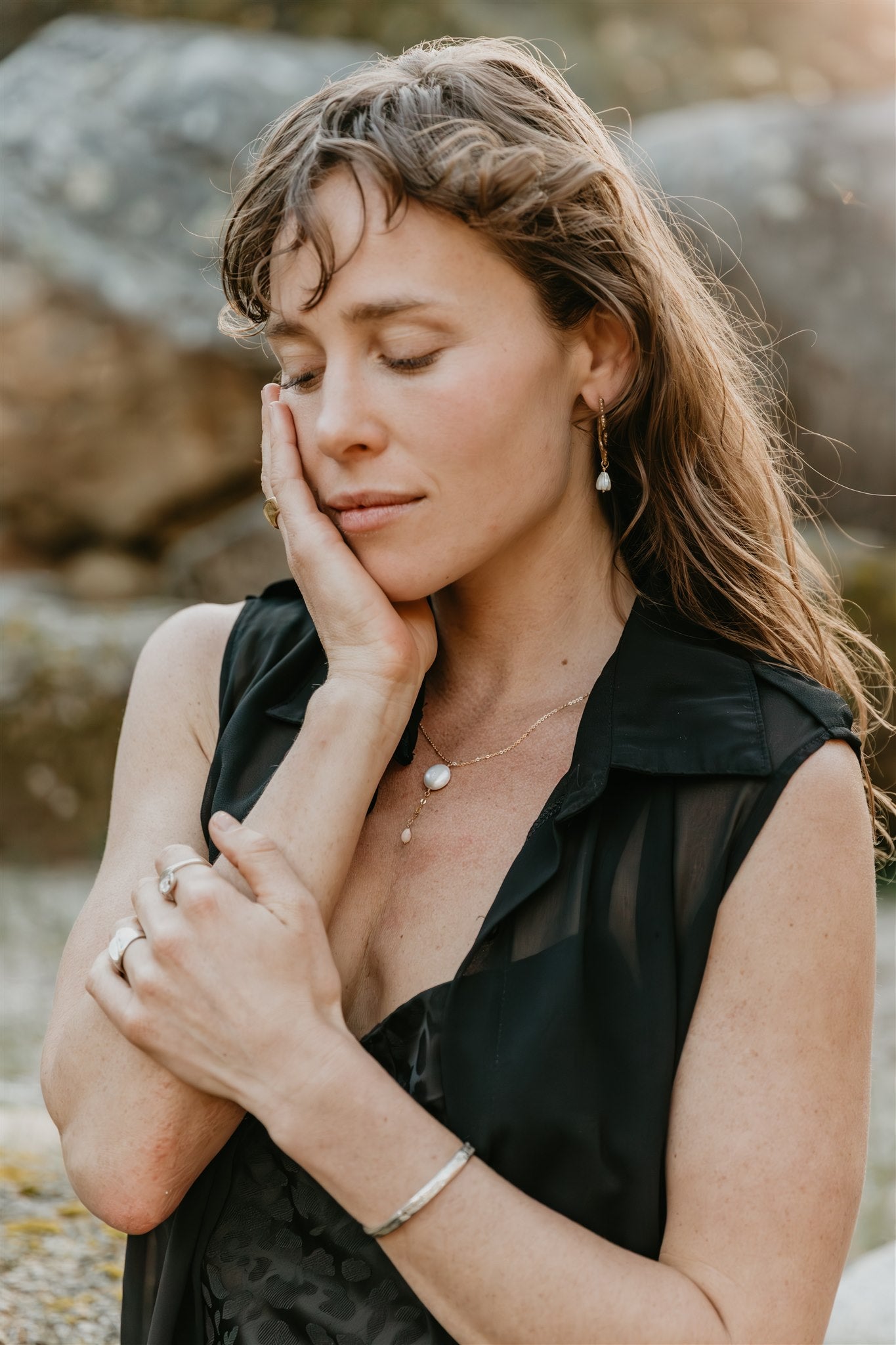 Woman in a black sleeveless top and lots of jewelry sitting outdoors with a blurred natural background