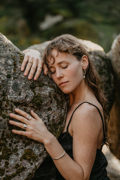 Woman in a black dress leaning against a rock with a blurred natural background, wearing jewelry