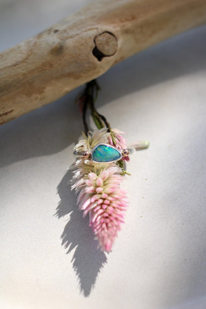 A ring with an Australian Boulder Opal, surrounded by rose gold accents, displayed on a pink flower on a neutral background.